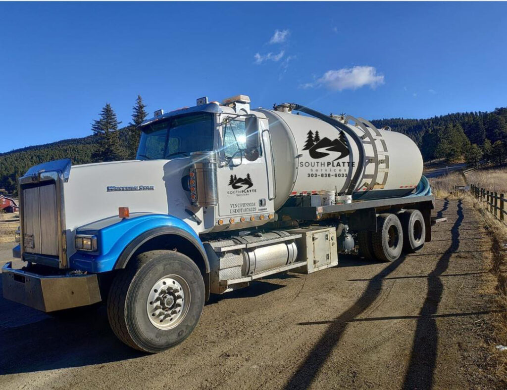 A white South Platte Services, LLC tanker and septic work truck with a view of mountains and forest behind it.